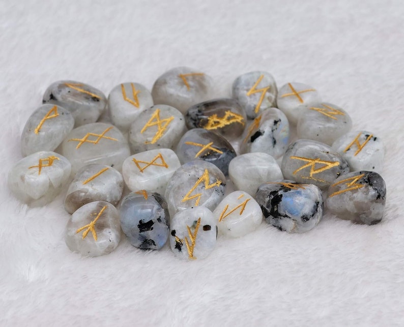 May include: A collection of polished, oval-shaped moonstone runes. Each stone is a mix of white, gray, and black with gold-colored etched symbols. The runes are arranged on a soft, white surface.