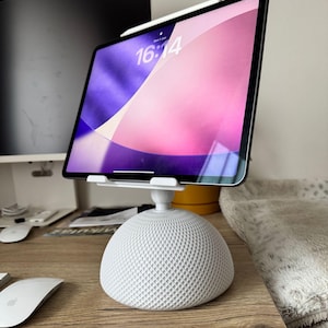 Peut inclure: Un support de tablette blanc en forme de dôme avec une surface texturée à motif quadrillé. Le support est doté d'une articulation centrale réglable soutenant une tablette dont l'écran affiche l'heure 16:14 et un fond abstrait violet et rose.