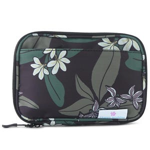 Puede incluir: Una bolsa de viaje rectangular con cremallera y estampado floral. El diseño presenta flores blancas y moradas, hojas verdes y un fondo oscuro. La bolsa tiene una cremallera negra y una pequeña etiqueta que dice "Happy Wahine."