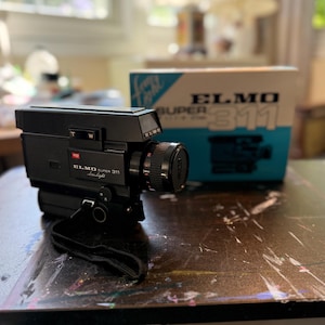 May include: A black Elmo Super 311 Low Light movie camera with a lens cap and a black wrist strap. The camera features a T and W zoom switch and is positioned in front of its original blue and white cardboard box labeled Elmo Super 311.