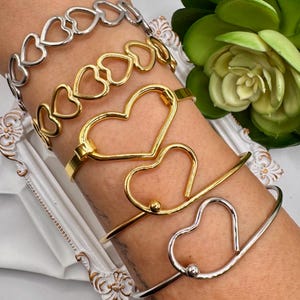 May include: A set of four gold and silver heart-shaped bracelets. The bracelets are stacked on a person's wrist. The bracelets are all different sizes and styles.