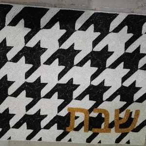 May include: A rectangular beaded mat with a black and white houndstooth pattern. The Hebrew word "Shabbat" is displayed in gold beads at the bottom. The mat is likely used for decorative or ceremonial purposes.