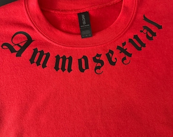 Może przedstawiać: Czerwona bluza z okrągłym dekoltem z napisem "Ammosexual" wydrukowanym czarnymi gotyckimi literami na piersi. Etykieta marki Gildan jest widoczna wewnątrz kołnierza.