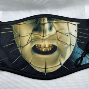 May include: Black face mask with a graphic print of a horror movie character with a face covered in pins and a wide open mouth with gold teeth.