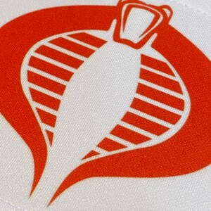 Może przedstawiać: Biała łatka z tkaniny z czerwonym logo kobry. Kobra patrzy wprost, ma otwartą paszczę i ma pasiasty wz&oacute;r na ciele.