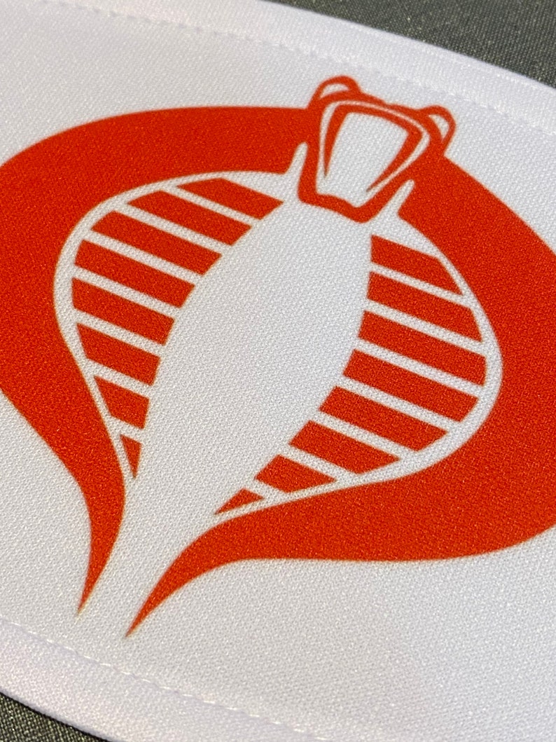 Może przedstawiać: Biała łatka z tkaniny z czerwonym logo kobry. Kobra patrzy wprost, ma otwartą paszczę i ma pasiasty wz&oacute;r na ciele.