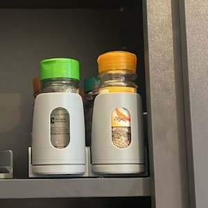 Puede incluir: Dos tarros de especias con tapas verde y naranja se almacenan en un estante de especias gris. Los tarros de especias son transparentes y contienen especias.