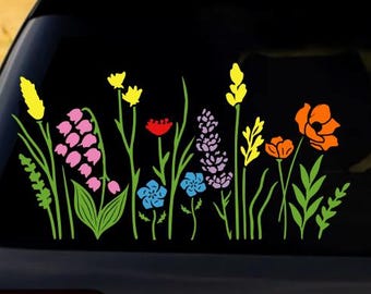 Op de afbeelding: Een kleurrijke bloemensticker met verschillende wilde bloemen in geel, roze, rood, blauw, paars en oranje. De sticker is aangebracht op de achterruit van een voertuig, met een vage buitenachtergrond.