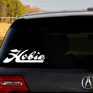 Op de afbeelding: Een witte vinyl sticker met het woord "Hobie" in een gestileerd lettertype is aangebracht op de achterruit van een zwarte SUV.