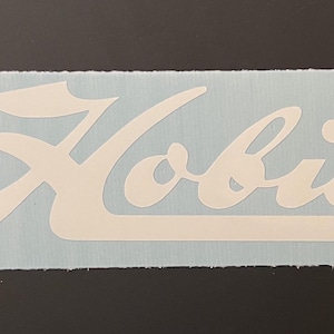 Op de afbeelding: Witte vinyl sticker met het woord "Hobie" in een gestileerd handschrift.