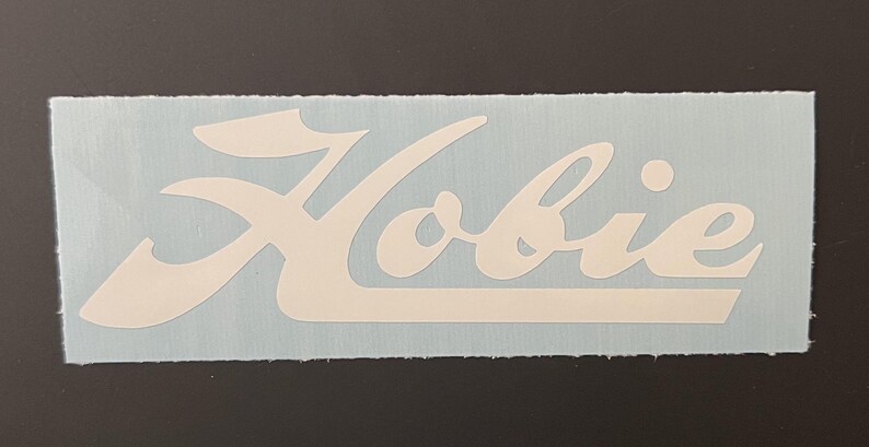 Op de afbeelding: Witte vinyl sticker met het woord "Hobie" in een gestileerd handschrift.