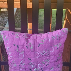 Op de afbeelding: Een roze gewatteerd kussen met een herhalend patroon van witte en groene ontwerpen. Het kussen is vierkant en heeft een knoop in het midden. Het rust op een blauw oppervlak.