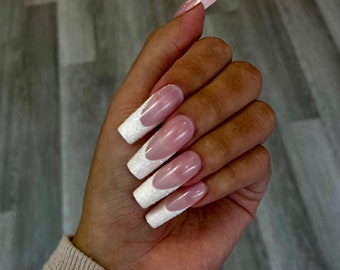 Könnte beinhalten: Eine Hand, die lange, quadratische Acrylnägel zeigt. Die Nägel haben ein klassisches French-Maniküre-Design mit rosa Basis und weißer, glitzernder Spitze. Die Nägel sind gut manikürt und poliert und zeigen einen modernen Nail-Art-Stil.