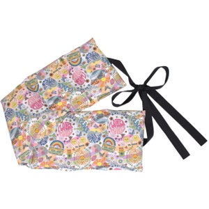 Könnte beinhalten: Ein langer, rechteckiger, weiß-rosa gemusterter Wärmekissen mit einer schwarzen Schleife. Das Muster zeigt Blumen, Schmetterlinge, Regenbögen und die Worte "Peace", "Flower Power" und "Here Comes The Sun".