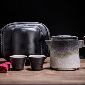 May include: A black travel case with a ceramic teapot and two teacups. The teapot has a wooden handle and a white and gray mountain design. The teacups are black with a white mountain design.