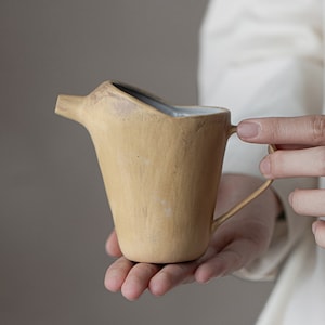 May include: A yellow ceramic jug with a distinctive, slightly curved spout and a handle. The jug has a light grey glaze on the inside.
