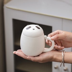 Può includere: Tazza in ceramica bianca a forma di panda con occhi neri e naso nero. La tazza ha un manico a forma di zampa di panda.