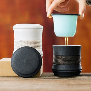 May include: Two ceramic teacups with lids. One is white with a brown rope detail and the other is black with a brown rope detail. A light blue tea infuser is on top of the black teacup.