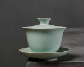 May include: A light green ceramic teacup with a lid and a matching saucer. The teacup and saucer have a subtle brown rim.