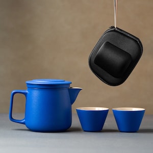 May include: A blue ceramic teapot with a lid and a matching blue ceramic teacup. The teapot has a spout and a handle. The teacup is sitting on a gray surface. A black carrying case hangs above the teapot and teacup.