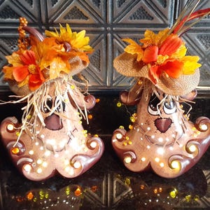 May include: Two brown ceramic gourds with painted faces and lights. Each gourd is decorated with a straw hat, fall leaves, and a flower.