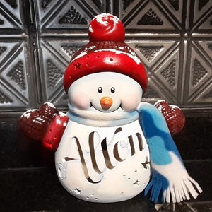 May include: A white ceramic snowman with a red hat and blue scarf. The snowman has a painted face with black eyes, a carrot nose, and a red smile. The snowman is holding a red and white mitten in each hand. The snowman has the word "Allon" written on its front.