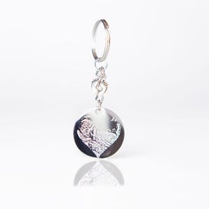 Op de afbeelding: Zilveren sleutelhanger met een ronde hanger met een gegraveerd hartontwerp. De hanger is bevestigd aan een zilveren ring en een zilveren ketting met een klein sieraad.