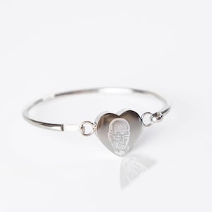 Könnte beinhalten: Ein silbernes Armband mit einem herzförmigen Anhänger, auf dem ein gravierter Abdruck einer Person mit Brille zu sehen ist. Das Armband ist aus Metall und hat ein schlichtes Design.