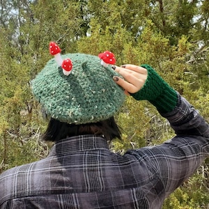 May include: A green crocheted beret with red and white mushroom embellishments. The beret is worn by a person with short black hair and a plaid shirt.
