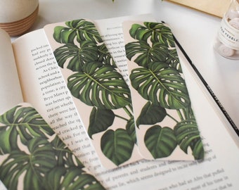 May include: Four white bookmarks featuring a green monstera leaf design. The bookmarks are placed on top of an open book with text.