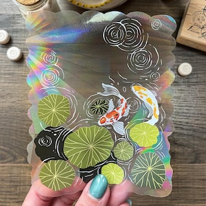 May include: Holographic sticker featuring a koi pond scene. The design includes two koi fish, lily pads, and water ripples. The sticker has a scalloped edge and iridescent finish, reflecting a rainbow of colors.