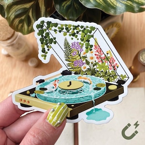May include: A sticker featuring a turntable design with a pond scene. The turntable's platter is a pond with koi fish, lily pads, and water flowing off the edge. The top of the turntable is a terrarium with flowers and greenery.