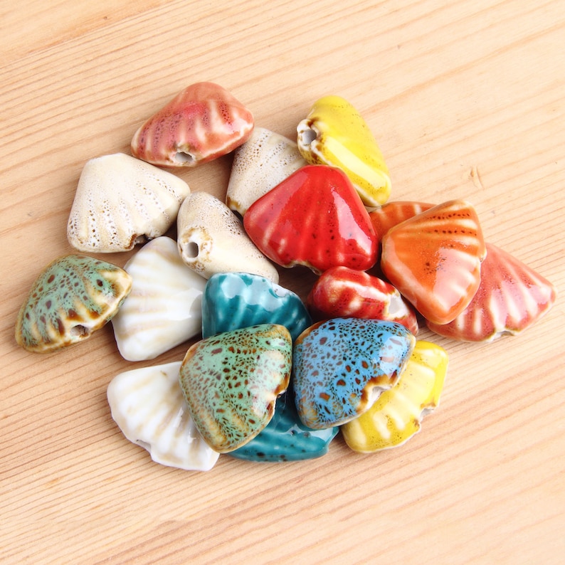 May include: Assortment of ceramic shell-shaped beads in various colors, including red, orange, yellow, blue, green, and white. The beads are glazed and have a speckled finish.