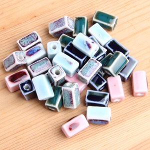 Könnte beinhalten: Eine Sammlung rechteckiger Keramikperlen in verschiedenen Farben, darunter Blau, Grün, Rosa und Weiß. Die Perlen haben eine glasierte Oberfläche und sind perfekt für die Schmuckherstellung.