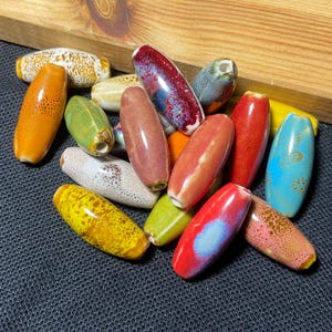 May include: A selection of brightly coloured ceramic beads in various shapes. The beads are mainly oval, with some leaf-shaped ones. Colours include orange, red, blue, yellow, green, and brown, with speckled and glazed finishes. They are on a dark surface.