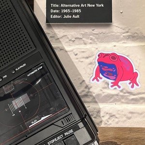 May include: A black cassette player with buttons labeled "Play", "REW", "F.F.", "Stop/Eject", and "Pause". The player is sitting on a wooden surface next to a white sticker of a pink and blue frog.