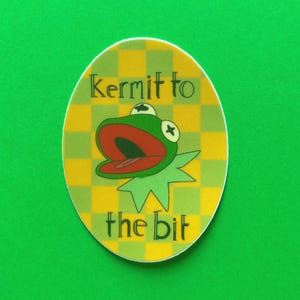 May include: Oval sticker featuring Kermit the Frog with an open mouth, set against a yellow and green checkered background. The text "Kermit to the bit" is printed on the sticker.
