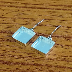 Może przedstawiać: Para srebrnych kwadratowych podstaw do kolczyków z zapięciem na haczyk. Podstawy są gotowe do wypełnienia żywicą lub innymi materiałami.