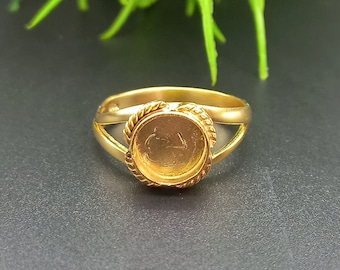Op de afbeelding: Een goudkleurige ring met een ronde zetting, met een gevlochten touwontwerp rond de rand van de zetting. De ring heeft een split-schacht ontwerp. De ring wordt gepresenteerd op een donkere ondergrond met groen gebladerte op de achtergrond.