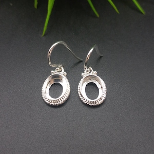 May include: A pair of silver-coloured earring findings. Each earring has an oval-shaped bezel setting with a decorative edge and a curved ear wire. The earrings are displayed against a dark background.