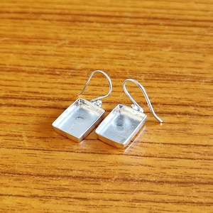 May include: A pair of silver-colored rectangular earring blanks with curved ear wires. The earring blanks have a recessed area for setting stones or other embellishments. The earrings are displayed on a wooden surface.