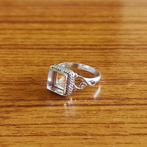 May include: A silver ring with a square bezel setting and a delicate, ornate design. The ring is on a wooden surface.