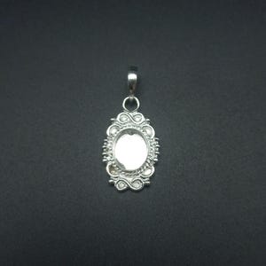 Può includere: Un ciondolo color argento con un'incastonatura di forma ovale. Il ciondolo presenta un bordo ornato, in stile filigrana, con volute decorative e dettagli a forma di perla. In alto c'è un piccolo anello per una catena.