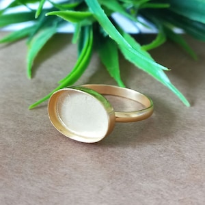 May include: A gold-toned ring with a round, blank, white bezel setting. The ring is on a brown surface.