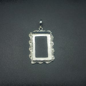 Puede incluir: Colgante rectangular plateado con un borde ornamental y enrollado. Tiene un lazo en la parte superior para una cadena y un centro oscuro y empotrado. El diseño es detallado y elegante, adecuado para la fabricación de joyas. Mide unos 3 cm.