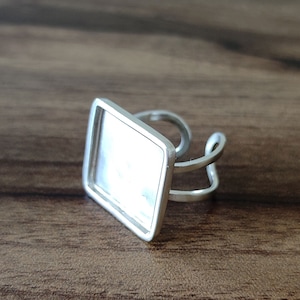 May include: A silver ring with a square-shaped bezel for a cameo or photo. The bezel is empty and ready for a custom design.