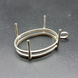 May include: A silver oval pendant setting with four vertical prongs and a small loop. The setting features a double-banded oval design, suitable for holding a gemstone or other decorative element. The setting is shown against a dark background.
