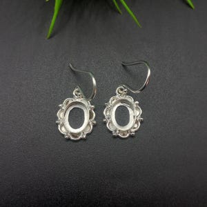 May include: A pair of silver earrings featuring an oval setting with a decorative, floral border. The earrings have a simple hook design. The background is a dark grey colour. The earrings are designed for jewellery making.