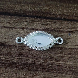 May include: Silver-toned metal pendant with a rounded oval shape and a beaded edge. The pendant has two small loops on either side for attaching a chain or cord.
