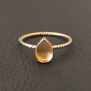 May include: A gold-toned ring with a twisted band and a teardrop-shaped bezel setting. The bezel is empty and ready to be filled with a gemstone.